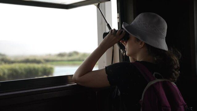 Woman birdwatcher observing birds from observation station in natural reserve. Female using binoculars for spotting rare birds in their natural habitat and environment. Standing at window and looking