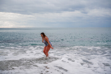 A plump woman in a bathing suit enters the water during the surf. Alone on the beach, Gray sky in the clouds, swimming in winter.