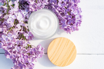 Obraz na płótnie Canvas Cosmetic cream with fresh branches of purple lilac blossoms on light wooden background. Dermatologic, components for cosmetics. Top view