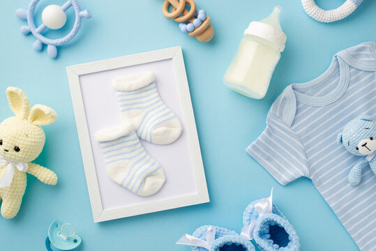 Baby accessories concept. Top view photo of photo frame with socks knitted bunny and bear toys blue shirt milk bottle teether rattle booties and soother on isolated pastel blue background