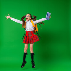happy schoolgirl in uniform and glasses, jumping in studio on green background. child with backpack...