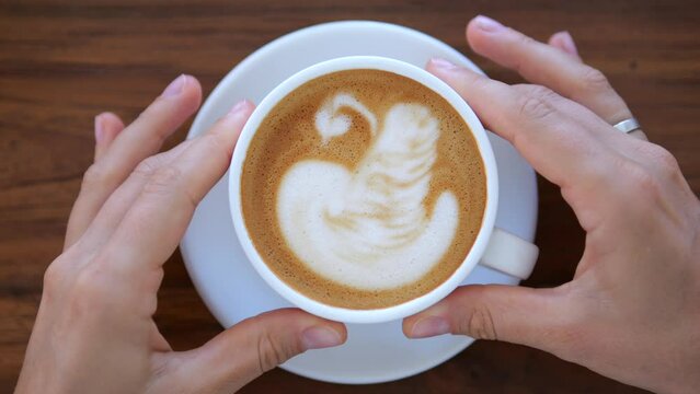 A mug of fragrant coffee held by female hands. Woman drinking morning sappuccino coffee from a white mug at a wooden table, top view. Coffee drink invigorates the body and spirit.