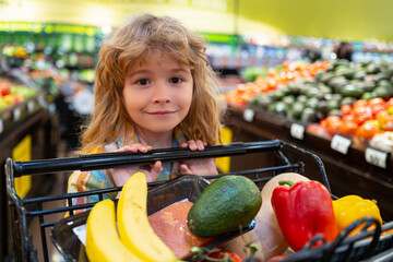 Healthy food for young family with kids. Portrait of smiling little child with shopping cart full of fresh vegetables. Kid having fun while choosing food in the supermarket.
