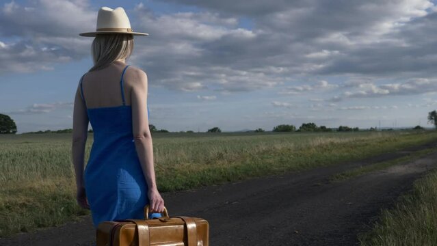 Girl in blue dress with suitcase on country road in summer.