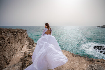 Happy freedom woman on the beach enjoying and posing in white dress. Rear view of a girl in a fluttering white dress in the wind. Holidays, holidays at sea.