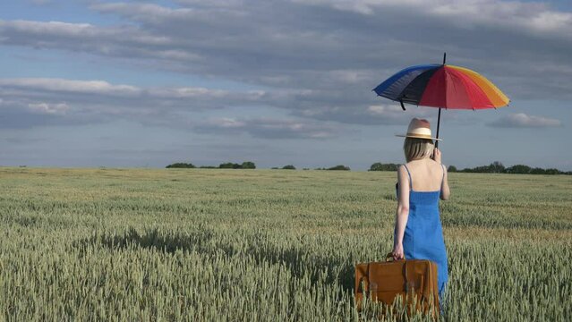 Girl in blue dress with suitcase and umbrella in wheat field