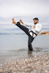 karateka martial arts expert in kimono performing a front kick on the shore of the beach splashing...