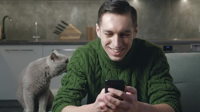 Smiling caucasian man in sweater sitting on sofa at cosy home with domestic cat and using smartphone surfing internet social media, watching videos