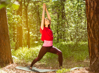 Woman doing yoga in park, standing in Virabhadrasana and looking up surrounded by lush greenery and trees