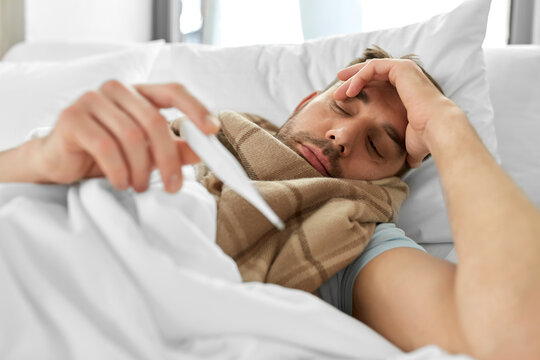 people, health and fever concept - sick man in scarf measuring temperature by thermometer lying in bed at home