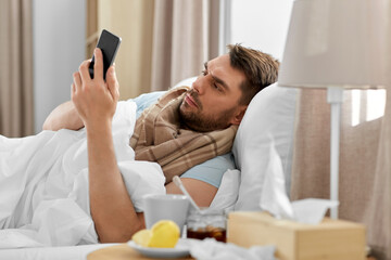 health, illness and people concept - sick man with smartphone in bed at home