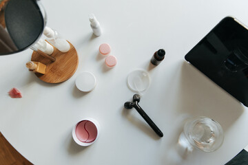 Top view of white table with cosmetic products, creams, patches, face massager, glass of water and planets. Wellness and self care concept