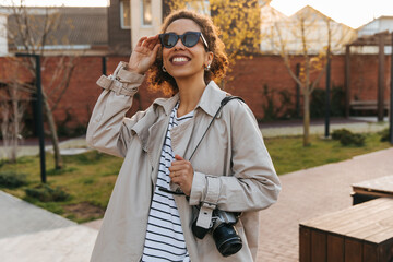 Fototapeta premium Positive young african woman admiring the autumn sky looking at it through sunglasses. Brunette with curly hair wears casual clothes. Lifestyle concept