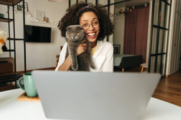 Happy young african woman holding cat in her arms chatting via video call on laptop. Brunette with curly hair wears casual clothes. Happy weekend concept.