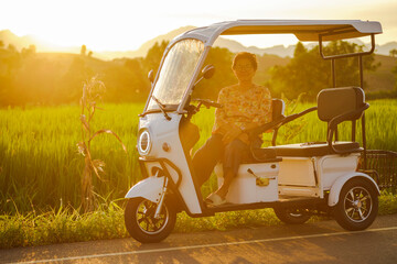 Smiling senior woman sitting on electric tricycle with rice fields and sunlight.