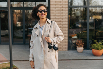 Beautiful young african girl holds camera, smiles, walks around city in good weather. Brunette with afro hair wears casual autumn clothes. Happy weekend concept.