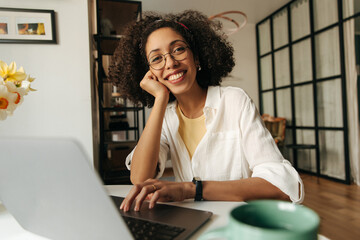 Happy young african woman looking at camera sitting at laptop home. Brunette with curly hair wears white shirt. Mood, lifestyle, concept. - obrazy, fototapety, plakaty