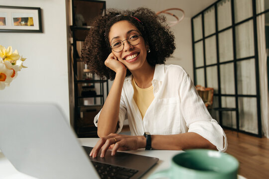 Happy young african woman looking at camera sitting at laptop home. Brunette with curly hair wears white shirt. Mood, lifestyle, concept.