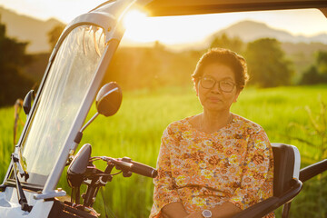 Smiling senior woman sitting on electric tricycle with rice fields and sunlight.