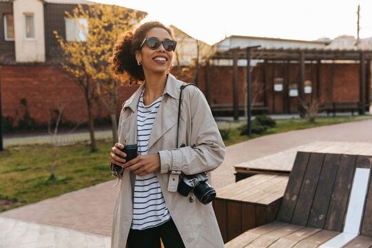 Pretty african young brunette girl walks around city carrying coffee, camera. Woman wears sunglasses, casual clothes. Rest time concept.