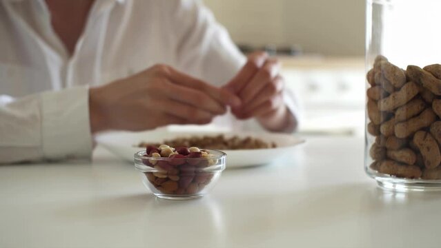 white young girl in a white shirt is sitting at table in the kitchen, peeling peanuts peeling off the peel of the nut Girl peeling raw peanuts for proper nutrition healthy breakfast high-calorie food
