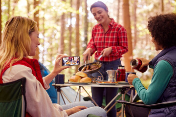 Woman Taking Photo Of Food As Female Friends On Camping Holiday In Forest Eat Meal Sitting By Tent