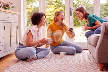 Group Of Female Friends Relaxing At On Sofa At Home Looking At Mobile Phone And Drinking Coffee