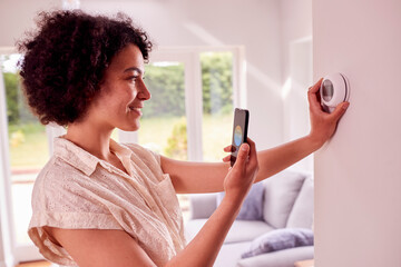 Woman Using App On Mobile Phone To Control Central Heating Thermostat At Home
