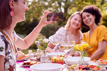 Woman Taking Photo Of Female Friends Sitting Outdoors In Garden At Home Eating Meal Together