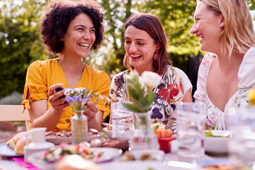 Three Female Friends Looking At Photos On Mobile Phone Eating Meal Outdoors In Summer Garden At Home