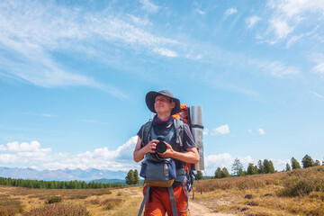 Fototapeta na wymiar Alone traveler with large backpack walks along hiking trail on sunlit high mountain plateau under white clouds in blue sky. Backpacker with photo camera in autumn mountain trekking in good weather.