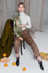 full length of stylish woman with flowers near vintage books and oranges on grey draped background.