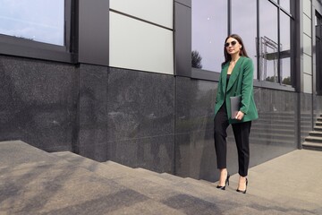 A young woman in a business suit with a laptop is walking near an office building