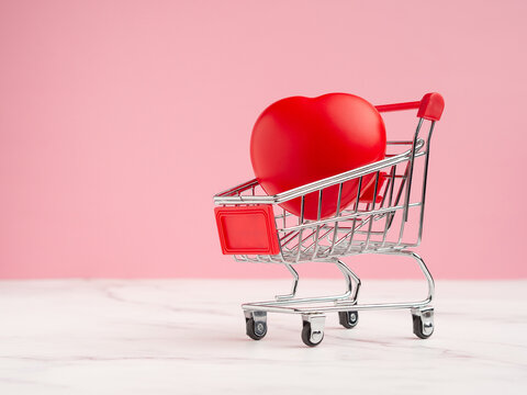 A mini shopping trolley with a red heart shape on a marble floor with a pink background