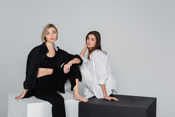 women in fashionable clothes sitting on black and white cubes isolated on grey.
