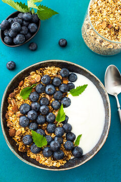 Homemade granola with yogurt, blueberries and mint leaves in a bowl on a turquoise background, delicious healthy breakfast