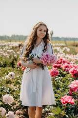 Happy young sexy girl standing in the middle of a flower field holding a peony bouquet. Single woman in white dress resting on pink background enjoying summer day. 