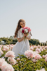 Fototapeta na wymiar Happy young sexy girl standing in the middle of a flower field holding a peony bouquet. Single woman in white dress resting on pink background enjoying summer day. 