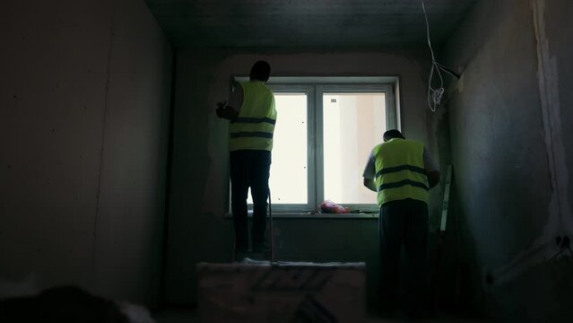 Silhouettes of plasterer painters applying plaster near the window of the room. Dark room and two silhouettes of workers making repairs in the room.