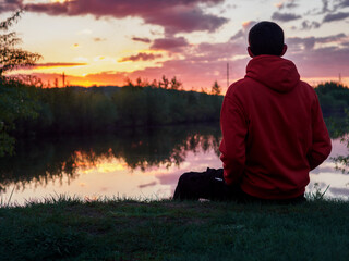 A young man is sitting on the riverbank at sunset, looking into the distance