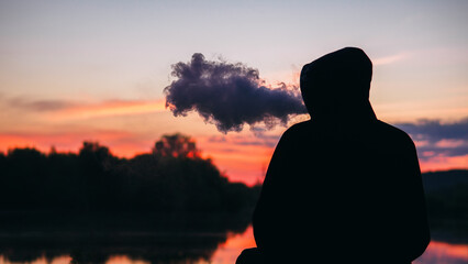 A man in a hoodie sits on the riverbank at sunset and exhales a cloud of steam with an electronic...