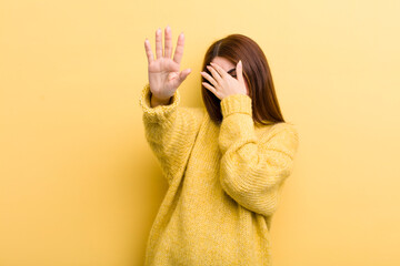 young adult pretty woman covering face with hand and putting other hand up front to stop camera,...