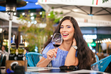 A young woman with bright makeup drinks wine from a glass of wine in a street cafe and laughs, looking at the camera
