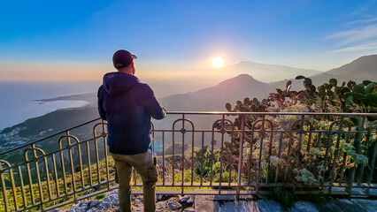 Tourist man watching beautiful sunset behind volcano Mount Etna near Castelmola, Taormina, Sicily,...