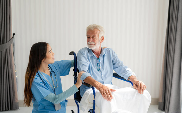 Friendly Staff Caregiver Of Nursing Home Talking To Elderly Man On Wheelchair In The Bedroom.