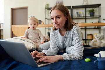 Young woman lying on bed and typing on laptop, she doing online work having a little child