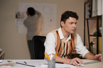 Concentrated architect working on computer when his colleague putting blueprint on wall in backg