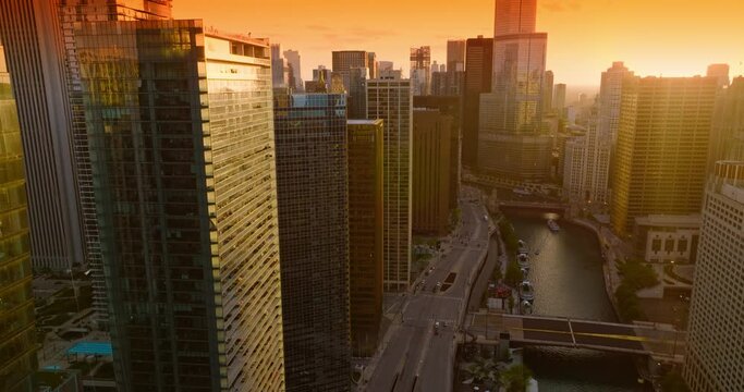 Beautiful setting sun reflecting in the mirror windows of skyscrapers. Drone flying along the roofs over Chicago River. Bright orange sky at backdrop.