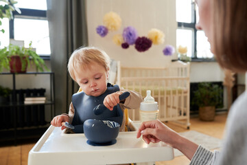 Little child in bib sitting in his chair and learning to eat porridge with spoon with the help of...
