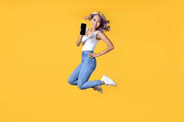 Fototapeta na wymiar Full length portrait of young Asian woman holding smartphone jumping on yellow background. Cheerful young female jumping up and showing mobile phone with empty screen, in studio